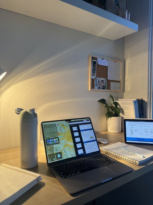 A cozy desk workspace featuring an open laptop, a water bottle, a tablet, a spiral notebook, and a potted plant, with warm sunlight casting a curved beam across the wall above a corkboard.