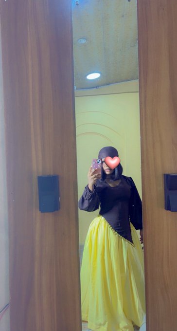 The image features a woman wearing a yellow dress with a black top and a red heart-shaped mask. She is taking a selfie using her cell phone while standing in front of a mirror, which has wooden doors behind her. The photo captures her reflection as she poses for the picture.