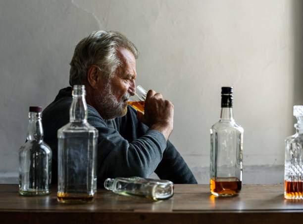 An older man with a gray beard sits at a wooden table surrounded by several empty liquor bottles, drinking from a glass.