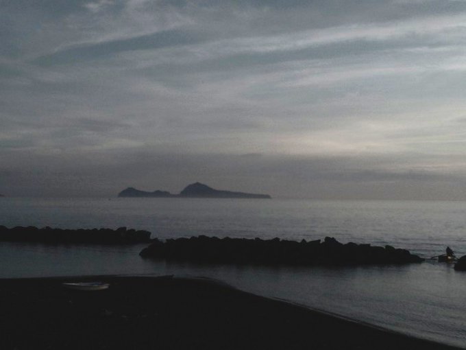 A serene seascape taken at dusk or dawn, showing a dark island silhouette in the distance, a rocky breakwater in the midground, and a calm beach in the foreground under a cloudy sky.