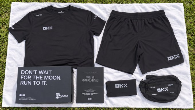 A black athletic kit (t-shirt, shorts, headband, fanny pack) and promotional materials laid out on a white sheet on grass, featuring the 'OKX' logo and the slogan 'DON'T WAIT FOR THE MOON. RUN TO IT.'