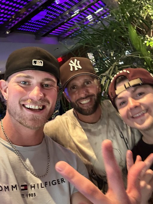 A selfie of three smiling young men wearing baseball caps, one with an MLB logo and another with a Yankees logo, posing closely together indoors.