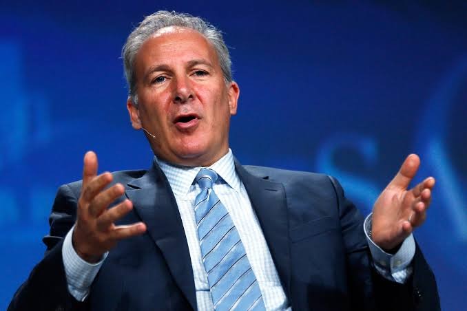 A professional photograph of a gray-haired businessman in a dark suit and blue striped tie speaking at a podium with a headset microphone, gesturing with his hands against a blue background.