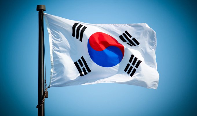 The South Korean national flag (Taegukgi) with its red-and-blue yin-yang center and black trigrams waving against a clear blue sky.