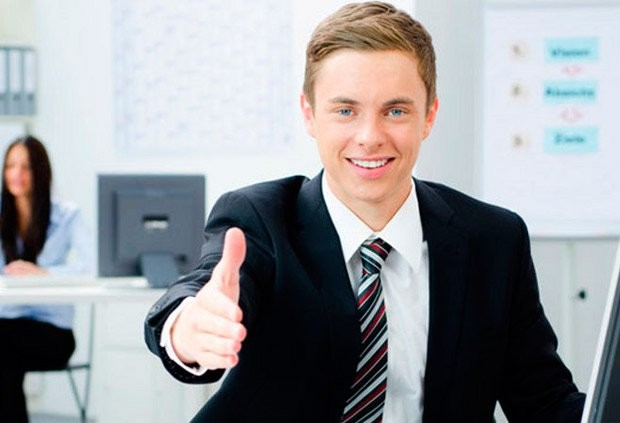 A smiling young businessman in a suit giving a thumbs-up in an office setting.