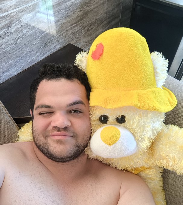 A man winks while posing shirtless next to a large yellow Care Bear plushie.