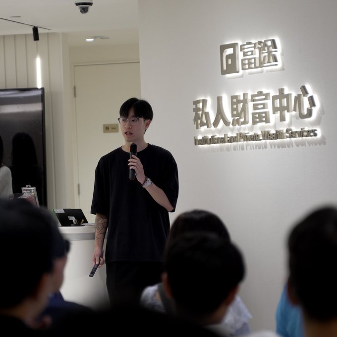 A man wearing glasses and a black t-shirt speaks into a microphone while presenting, with the logo '富途 私人財富中心 (Institutional and Private Wealth Services)' displayed on the wall behind him to an audience.
