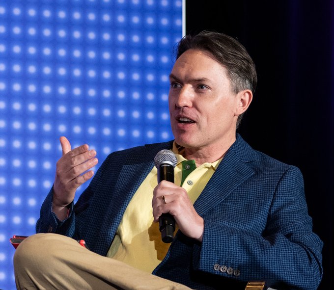 A man in a blue checkered blazer and yellow shirt speaks into a microphone while gesturing, seated against a blue dotted backdrop that suggests a panel or conference setting—likely capturing a moment from his time at Blockworks before his departure.