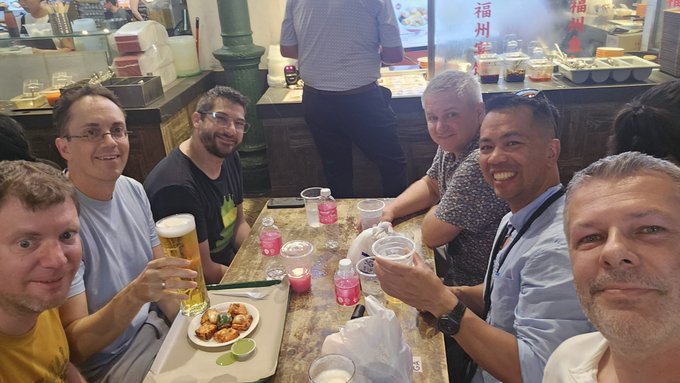 Group of six men smiling at a restaurant table, with drinks, food, and Chinese signage '福州' visible in the background.