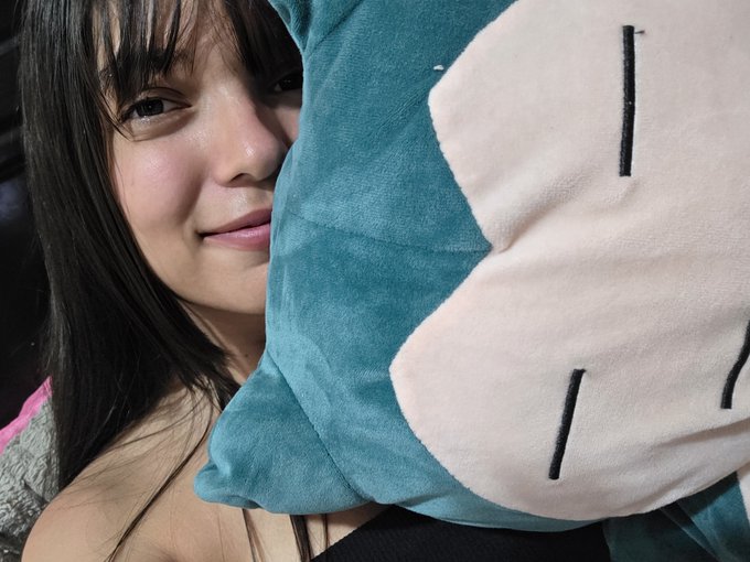 Close-up selfie of a smiling young woman with dark hair holding a teal plush toy with a white face design next to her cheek.