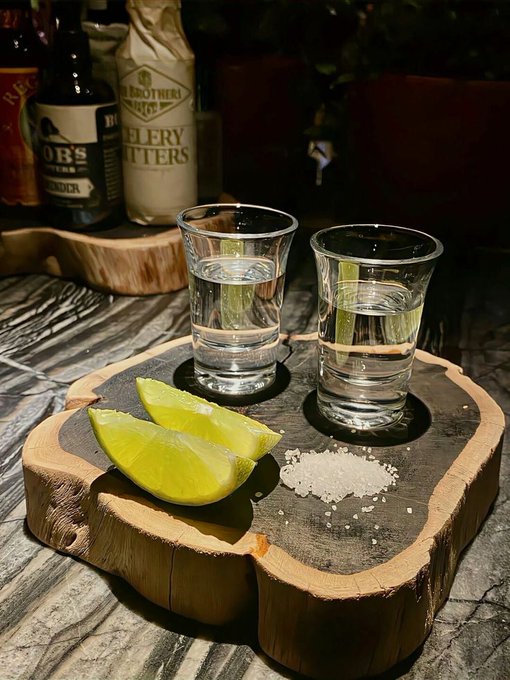 Two shot glasses with clear liquid sit on a wooden serving board alongside lime wedges and a pile of salt, with liquor bottles including O'B's and 2 Brothers Brewery visible in the background.