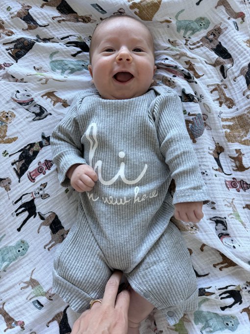 A smiling baby wearing a gray ribbed onesie with white text, lying on a white blanket patterned with various dog illustrations.