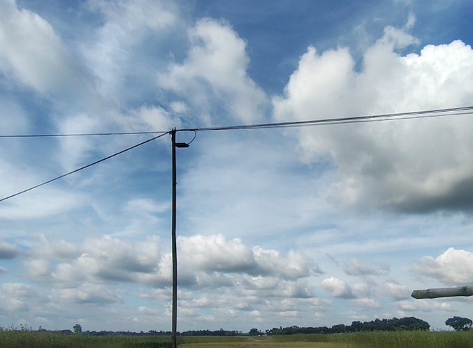 A landscape view featuring a wooden utility pole with power lines against a bright blue sky filled with fluffy white clouds over a grassy field.