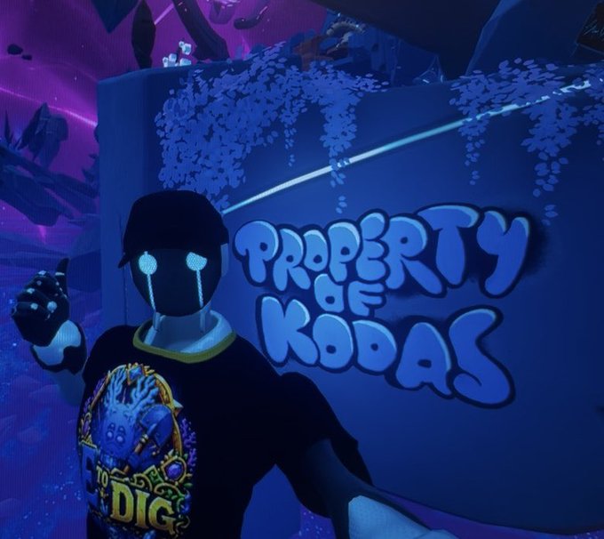 A virtual character wearing a blue "I DIG" t-shirt and glowing teary eyes stands in front of a large blue wall displaying the text "PROPERTY OF KODAS" in bubble letters, likely showcasing the new merch referenced from the Nexus market in OthersideMeta.