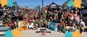 A large group of people pose for a photo outdoors at a paintball event, with a banner reading 'BORED PAINTBALL CLUB EST 2020'.