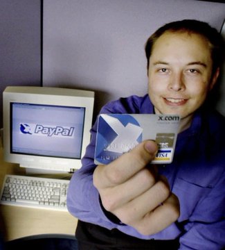 An archival photo of a young man smiling while holding up two credit cards (PayPal and X.com branded), seated in front of an early desktop computer displaying the PayPal logo.