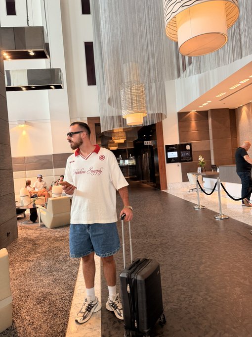 A man wearing sunglasses, a "Supreme Supply" jersey, denim shorts, and sneakers stands with a rolling suitcase in a modern hotel lobby, ready to start the day. The casual "Gm friends" greeting pairs with his relaxed travel-ready look as he appears to be heading out or checking in.