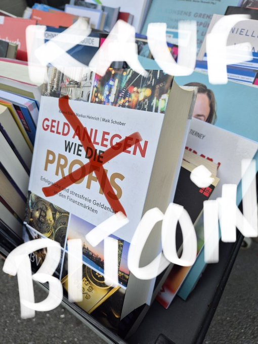 A display of books at an outdoor stand with a German book titled 'GELD ANLEGEN WIE DIE PROFIS' crossed out in red paint, while the words 'KAUF' and 'BITCOIN' are written in white paint on the glass.