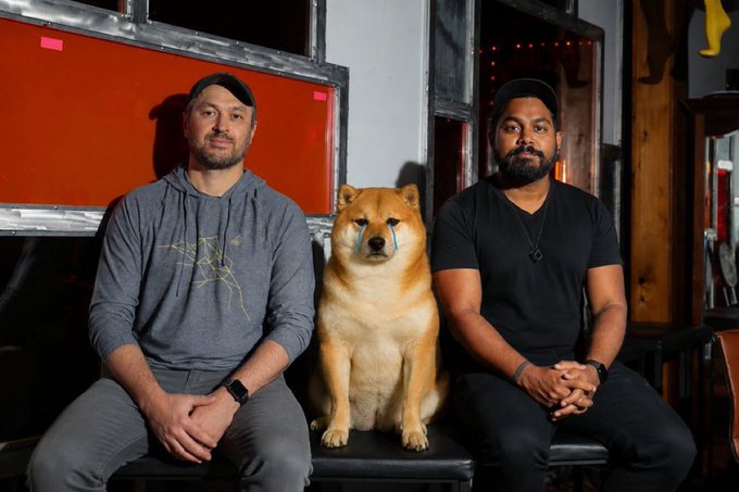 Two men in black caps sitting side by side with a Shiba Inu dog between them in a studio with red panels.