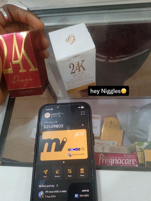 A first-person view of a hand holding perfume boxes ('Rouge' and '24K Elixir') next to a smartphone displaying a crypto wallet with a balance of '~$21.09802' and an 'Off-ramp USDC to NGN' transaction history.