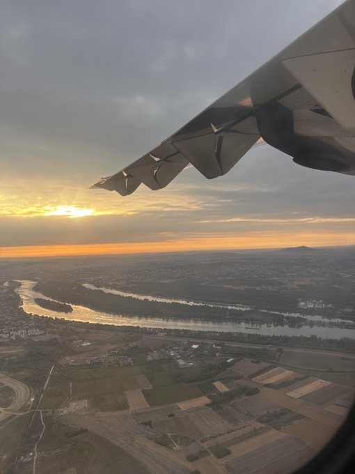 Aerial view from an airplane window showing the wing of the aircraft against a sunset sky, with a winding river, patchwork farmland, and distant mountains visible below.