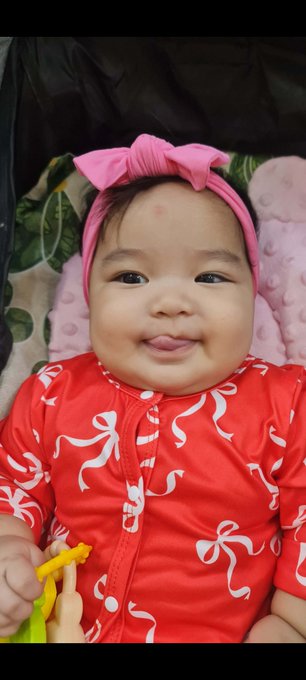 A baby with a pink bow headband and a red patterned outfit smiles and sticks out their tongue playfully while holding a yellow toy. The image accompanies a post sharing a message about God's grace alongside a link to an NFT collection.
