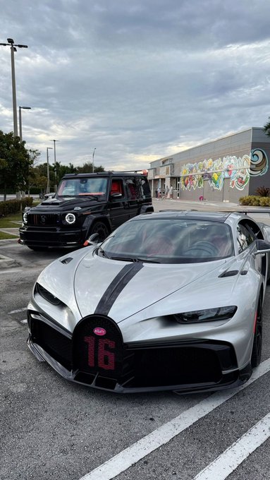 A silver Bugatti hypercar with a black stripe and the number '16' on its front, parked next to a black Mercedes-Benz G-Wagon in a parking lot with a mural visible in the background.