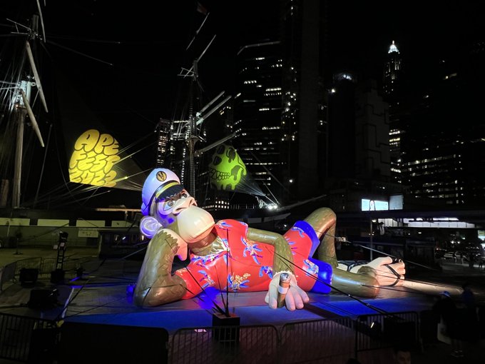 A large illuminated inflatable sculpture of a relaxed figure in a Hawaiian shirt and swim trunks reclines on a platform at night, set against a backdrop of city skyscrapers and a tall ship.
