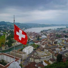 A large Swiss flag flies above Bern, overlooking the city and a lake. The image illustrates the accompanying news that Swiss regulator FINMA approved the country’s first retail Bitcoin ETF.