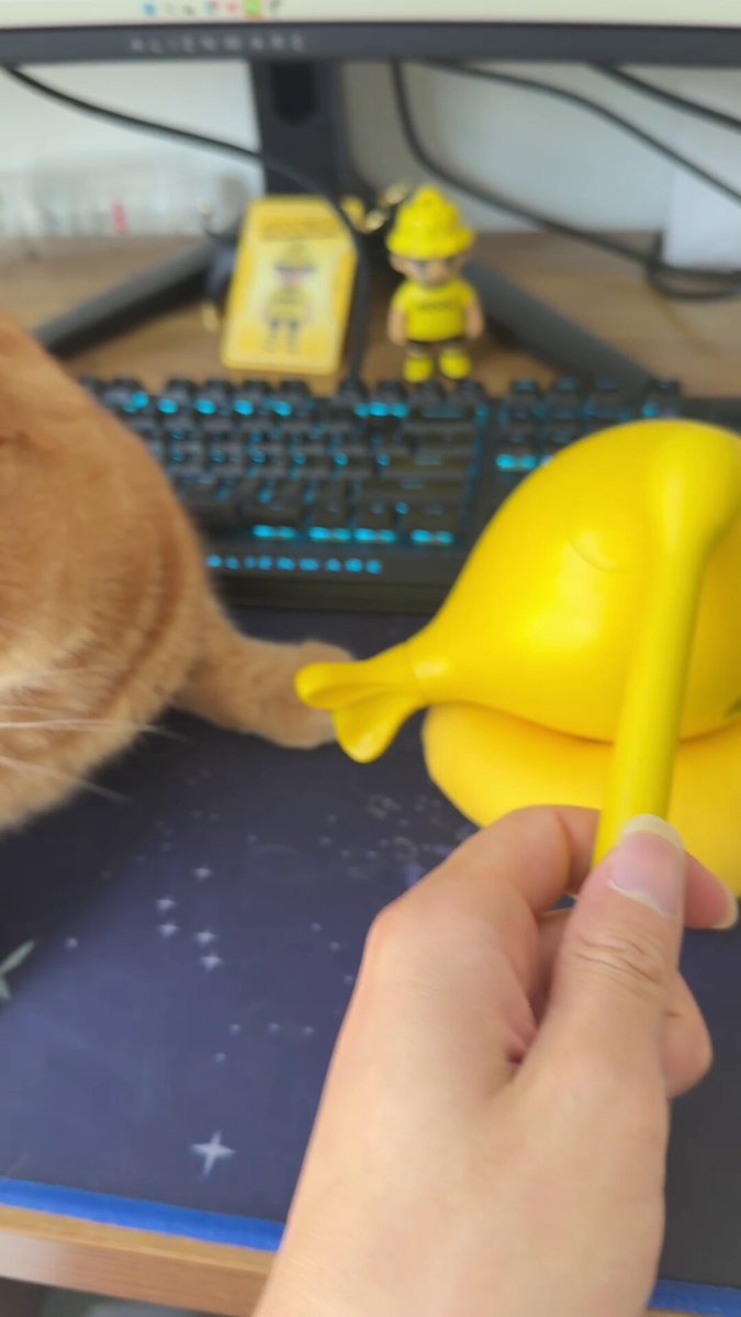 The image features an orange cat sitting next to a computer keyboard with a yellow plastic fish toy placed on top of it. The cat appears to be playing with or interacting with the toy, while the keyboard remains unoccupied.
