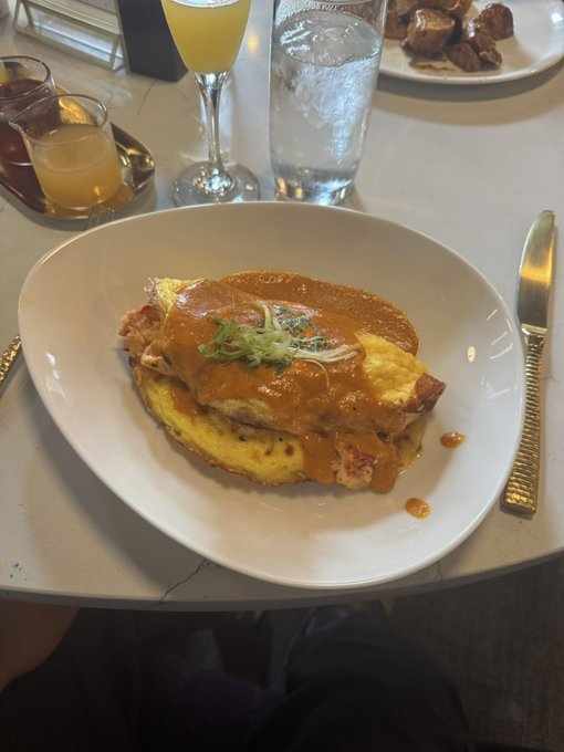 A plated meal of what appears to be eggs Benedict or a similar dish with rich orange sauce and green garnish, served on a white plate alongside a mimosa and other beverages at a restaurant.