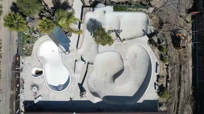 An aerial view of a concrete skatepark under construction, featuring curved bowls and ramps surrounded by palm trees and construction equipment.