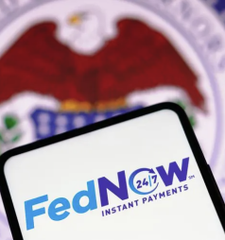 A smartphone displaying the FedNow logo with '24/7 INSTANT PAYMENTS' text, with a blurred government seal in the background.