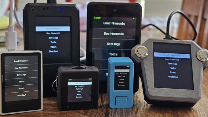 Several cryptocurrency hardware wallets are displayed on a wooden table, all showing the same menu screen with options including "Load Mnemonic," "New Mnemonic," "Settings," "Tools," "About," and "Shutdown." The devices vary in size and design, with one device in the center labeled "test" at the top, illustrating the post's point about verifying open-source firmware across different hardware wallet platforms.