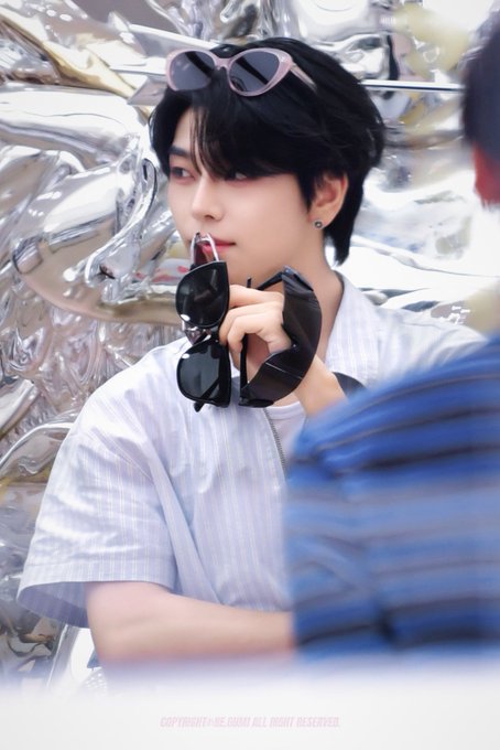 A young man with dark hair poses stylishly with pink sunglasses perched on his head while holding black sunglasses in his hand, photographed at the "260723 Double Lovers Pop-up #HeXinchengLong" event, accompanied by another person in a blue striped shirt.