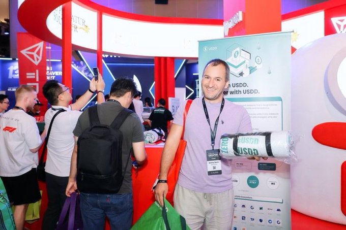A man smiling at a trade show or convention, holding USDD branded merchandise, with a USDD promotional banner visible in the background.