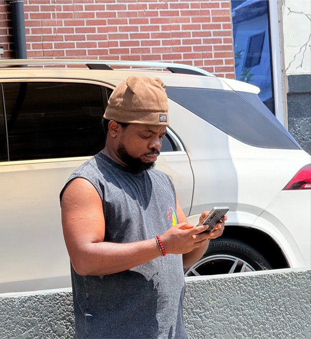 A man in a gray sleeveless shirt and brown beanie stands in front of a white SUV, focused on his smartphone, seemingly unaware of the cautionary crypto message warning about the "67" token scam.

The partially visible tire displays text including "TURANZA" along with numbers, while a small logo patch is visible on his beanie.
