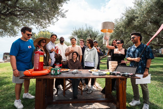The image features a group of people gathered around an outdoor table with various objects on it, including a red fire extinguisher, a lamp, and some books. The people are posing for a photo, likely celebrating LI.FI First Things First's recent success in shipping their architectural solution to the market.