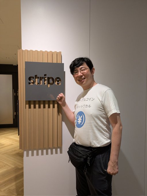 A man with glasses wearing a white t-shirt with a blue yen symbol and Japanese text stands next to a 'stripe' sign in Japanese.
