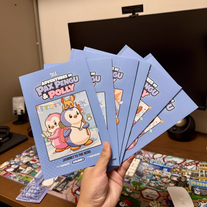 A hand holds a fanned-out stack of five blue booklets titled 'Adventures of Pax Pengu & Polly - Volume 1: Journey to the Berg,' featuring cartoon penguin characters on the cover.