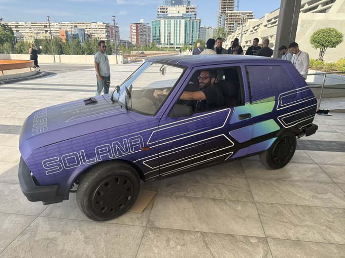 A small purple compact car with 'SOLANA' branding is parked on a tiled plaza, with a bearded driver inside and onlookers standing nearby.
