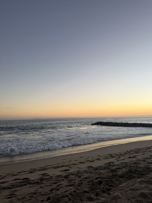A serene beach scene at sunset or sunrise, with gentle waves rolling onto a sandy shore marked by footprints, under a sky transitioning from soft blue to warm golden hues near the horizon.