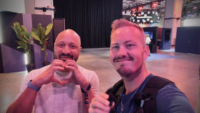 Two smiling men pose at what appears to be a convention or trade show; the bald man on the left forms a heart with his hands while the bearded man on the right gives a thumbs up.