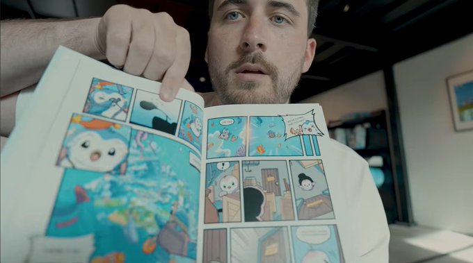 A man with a beard holds up an open comic book or manga, revealing colorful panels with cute characters and ocean scenes.