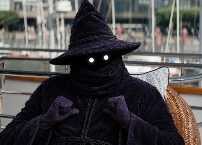 A figure dressed in a dark purple hooded robe with glowing white eyes sits on a wicker chair, evoking a sorcerer or mystical character.