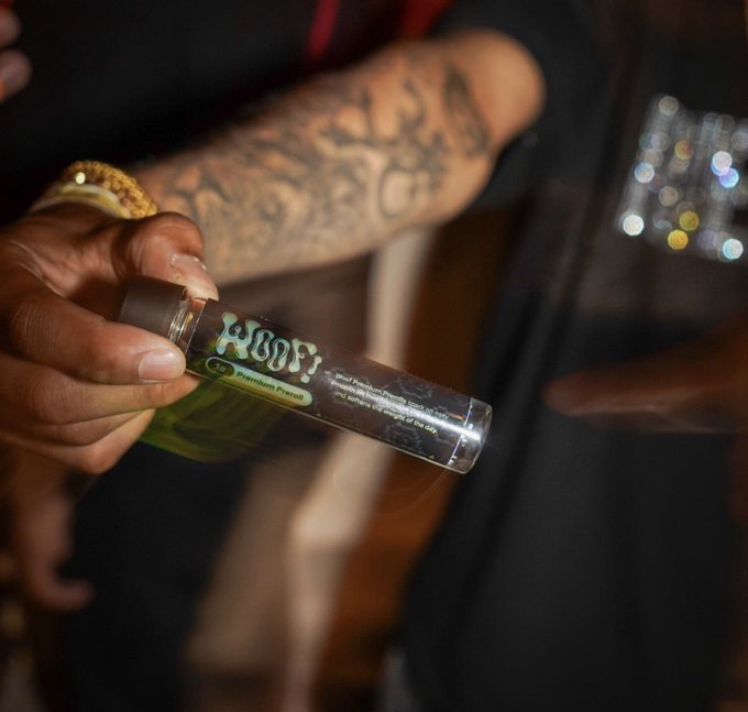 A close-up shot of tattooed hands holding a clear glass tube containing green liquid, labeled 'HOOD! by Premium Loud' cannabis product.