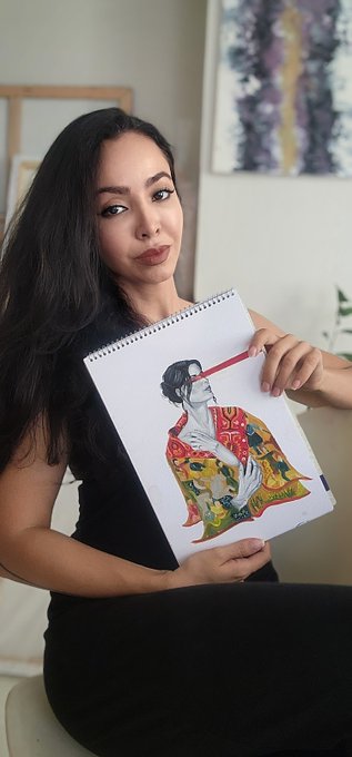 A young woman with long dark hair, wearing a black dress, sitting indoors and holding up a sketchbook displaying an artistic illustration of a figure with comic-book style panels.
