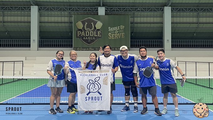 A group photo of seven people posing on an indoor pickleball court at Paddle Ranch, wearing blue 'Ronin' shirts and holding paddles, with one person holding a white banner reading 'Sprout Pickleball Club' featuring a bunny logo, while a 'Paddle Ranch' banner reading 'Saddle Up & Serve' hangs in the background.