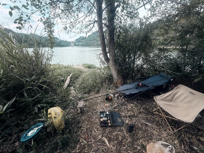 A serene lakeside camping setup featuring a camping chair, fishing rods, tackle box, and a kayak on the grassy shore with mountains in the background.