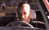 The image shows a bald man sitting inside a car, looking out of the window with his eyes closed. The car appears to be parked or stationary, as there are no other vehicles visible in the scene. The focus of the image seems to be on the man and his surroundings rather than any specific details about the car's interior or exterior features.